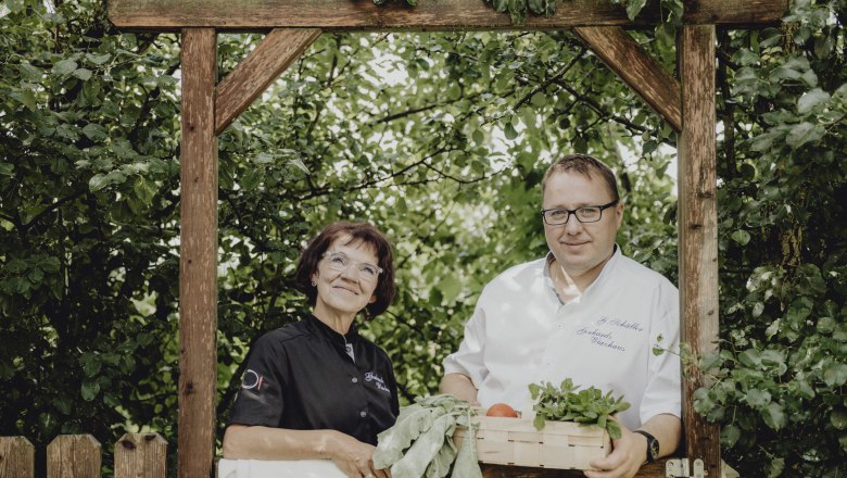 Wirtsleute Gerhard und Bernadette Schäller, © Niederösterreich Werbung/Rita Newman Zwei Personen stehen unter einem Holzbogen im Garten, eine Frau in schwarzer Kleidung und ein Mann in weißem Hemd mit einem Korb voller Gemüse.