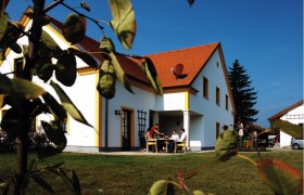 Ferienwohnungen Weixelbaum, © Ferienwohnungen Weixelbaum Ein weißes Haus mit rotem Dach, umgeben von grünem Rasen und Bäumen. Menschen sitzen an einem Tisch im Freien.