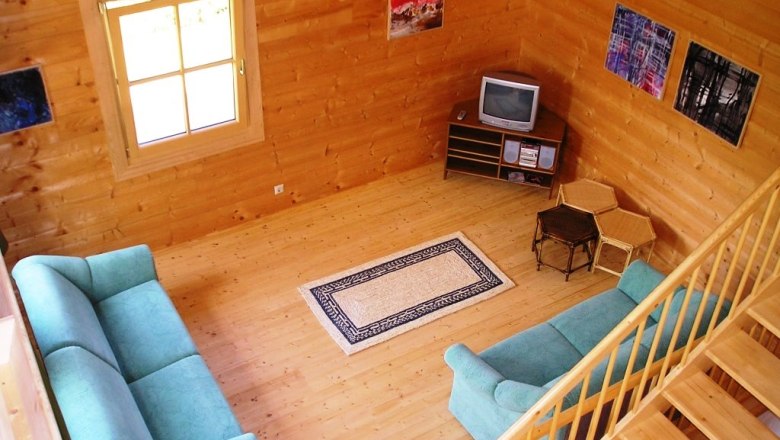 Wohnzimmer, © Daniel Fries Holzvertäfeltes Wohnzimmer mit zwei grünen Sofas, einem kleinen Fernseher und einem Teppich auf Holzboden.