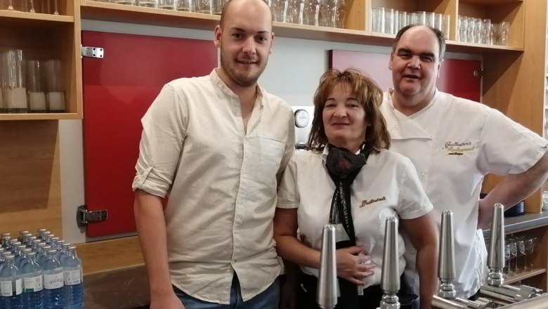 Wirtsfamilie Vater, Mutter, Sohn hinter Tresen, © Gasthubers Wirtshaus Drei Personen stehen hinter einer Theke mit Zapfhähnen und Gläsern im Hintergrund.