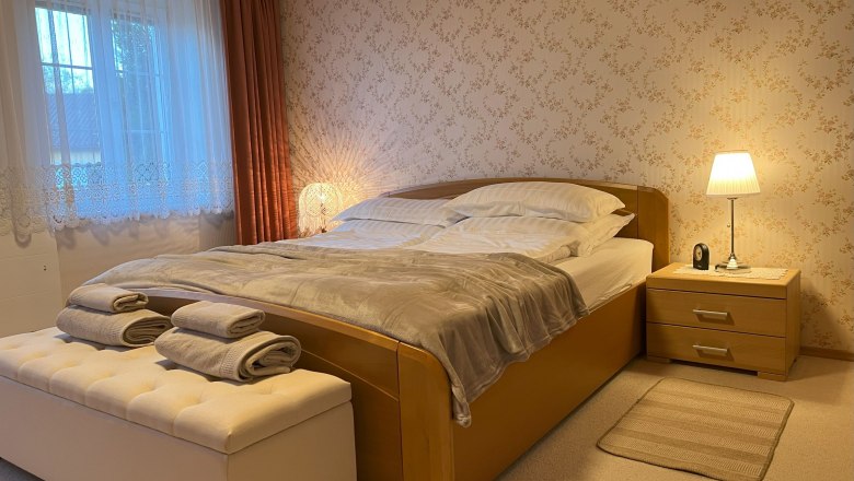 Schlafzimmer, © Waltraud Forstner Gemütliches Schlafzimmer mit Doppelbett, Nachttischlampe und Fenster mit Vorhängen.