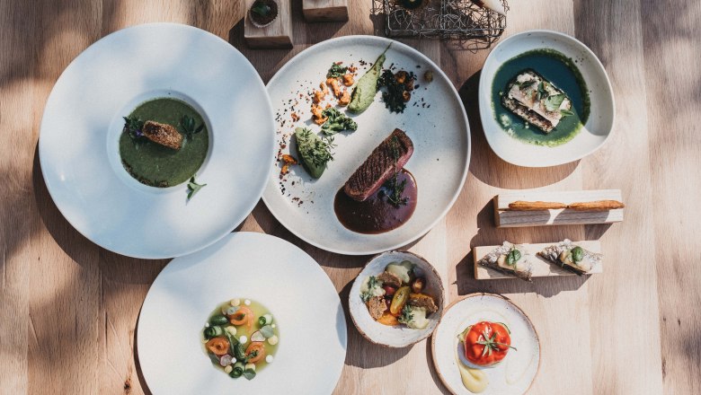 Köstliches aus der Haubenküche, © Molzbachhof Verschiedene Gourmetgerichte auf weißen Tellern, kunstvoll angerichtet auf einem Holztisch.