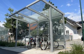 E-Bike Ladestation Haus der Digitalisierung, © Stadtgemeinde Tulln, Manuel Hofbauer E-Bike-Ladestation mit überdachtem Bereich und Fahrrädern, im Hintergrund ein Haus mit Solarpanelen.