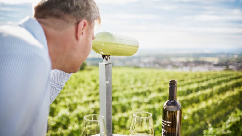 Weinweg Langenlois, © © Waldviertel Tourismus, Gerhard Wasserbauer Ein Mann blickt durch ein Fernglas auf ein Weinfeld. Auf dem Tisch stehen eine Weinflasche und zwei Gläser.