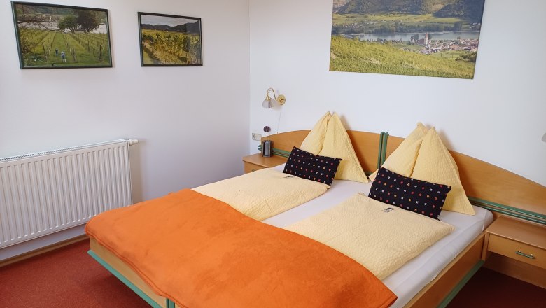 Haus Barbara_Zimmerbeispiel, © Haus Barbara Gemütliches Hotelzimmer mit Doppelbett, orangefarbener Decke und Landschaftsbildern an der Wand.