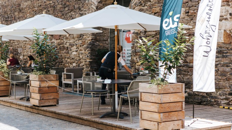 Café Wohnzimmer Außenansicht, © FMS Steinertor Gastronomie BetriebsgmbH Außenbereich eines Cafés mit Sonnenschirmen und Holzkisten als Pflanzgefäße vor einer Steinmauer.