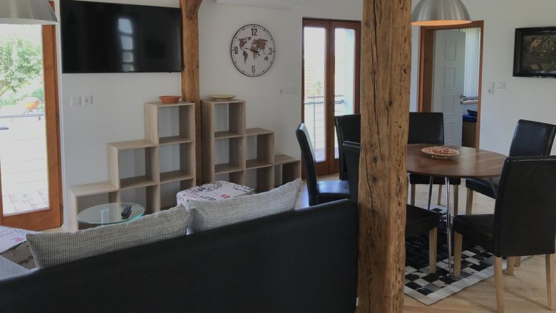 Wohnbereich, © Familie Mokesch Moderner Wohnbereich mit Holzbalken, Esstisch, Stühlen, Wandregal, Uhr und Fernseher.