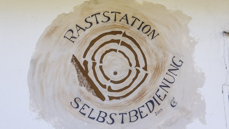 Radlerrast, © Fam Wingelhofer Krimmel Wandgemälde mit der Aufschrift 'Raststation Selbstbedienung' und einem stilisierten Baumstamm.