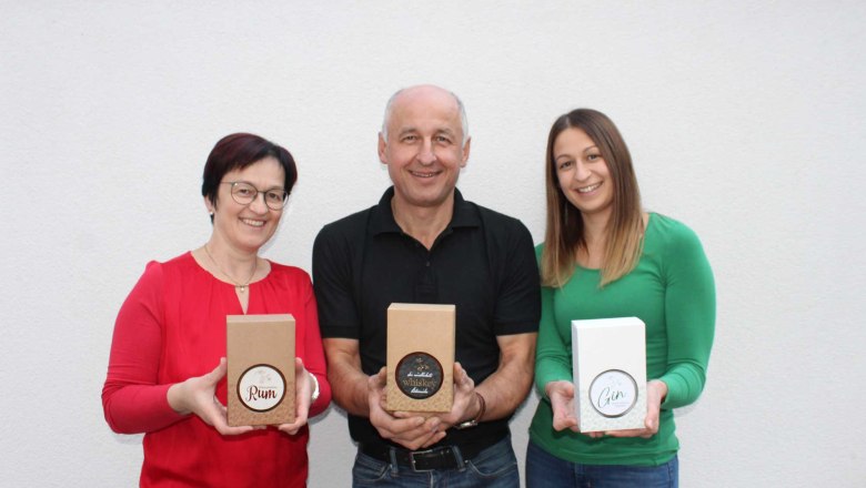 Familie Kainz, © Heuriger Kainz Drei Personen halten Geschenkboxen mit Rum, Whiskey und Gin vor einer weißen Wand.