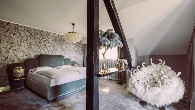 Hochzeitssuite, © Matthias Kronfuss Luxuriöses Schlafzimmer mit großem Bett, dekorativer Tapete, Kronleuchter und extravagantem Sessel mit Schwanendesign.
