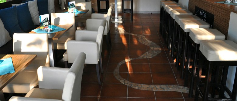 Carpe Diem_Innenansicht, © Carpe Diem Innenansicht eines Cafés mit weißen Stühlen und Tischen, braunem Fliesenboden und Barhockern.