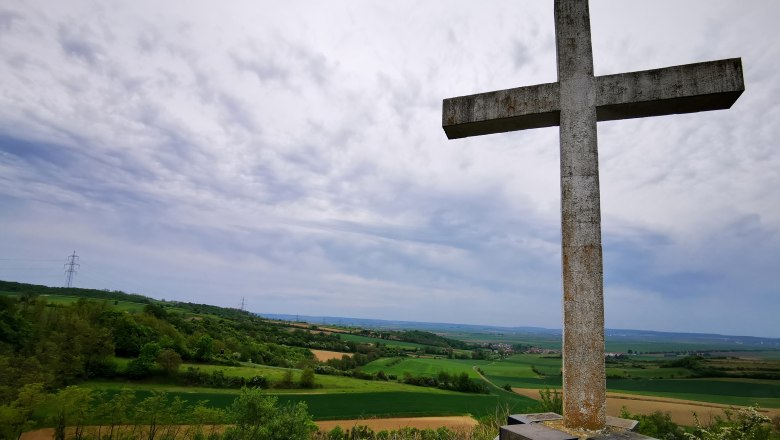 Kogelkreuz, © Weinstraße Weinviertel Ein großes Steinkreuz auf einem Hügel mit Blick auf eine grüne Landschaft und bewölkten Himmel.