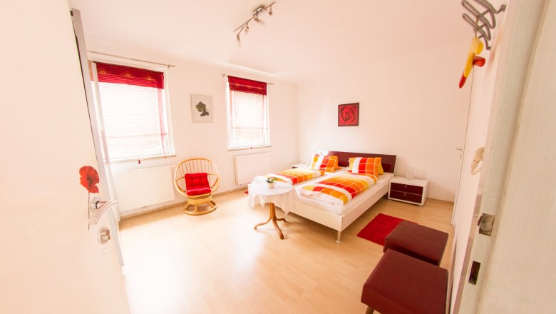 Gästezimmer Winzerhof Lackinger, © Kastner New Media Helles Gästezimmer mit Doppelbett, roten und gelben Bettdecken, zwei Fenstern, einem runden Sessel und einem kleinen Tisch.