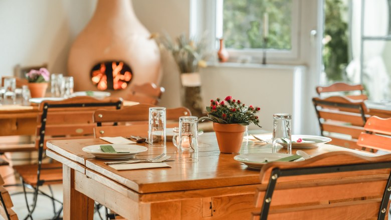 Frühstücksraum, © Matthias Auer Ein gemütlicher Frühstücksraum mit Holztischen und -stühlen, gedeckt mit Geschirr und Gläsern. Ein Kamin im Hintergrund sorgt für eine warme Atmosphäre.