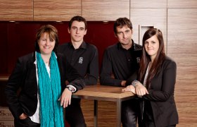 Familie Schagl, © Weingut Schagl Vier Personen stehen lächelnd vor einer Holzwand.