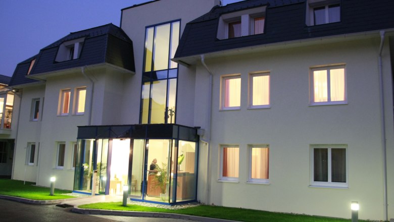 Hoteleinfahrt, © City Hotel Mödling Eingangsbereich eines modernen Hotels bei Dämmerung mit beleuchteten Fenstern und Glasfassade.