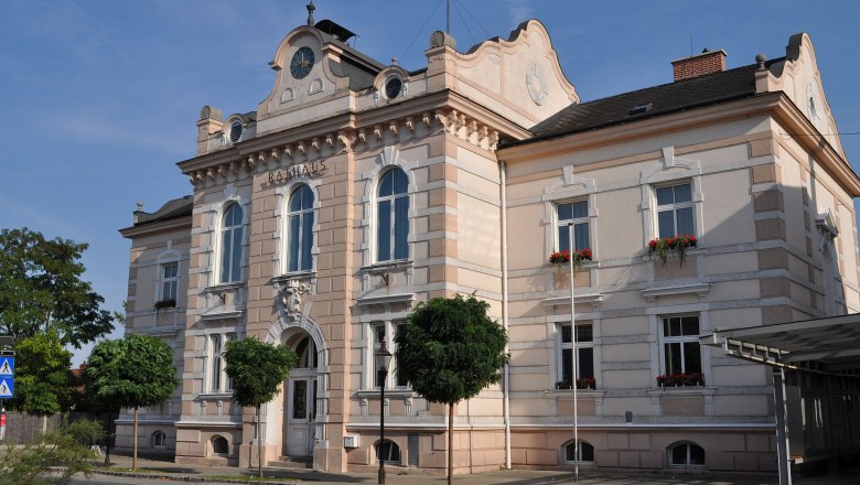Rathaus Felixdorf, © Spiderman183 Das Rathaus von Felixdorf, ein historisches Gebäude mit Uhrturm und verzierten Fenstern, umgeben von Bäumen und einer Straße im Vordergrund.