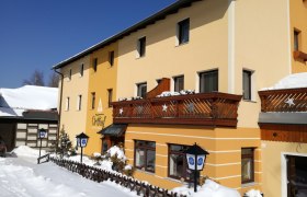 Hausansicht, © Maria Kager Gelbes Gebäude mit Holzbalkonen im Schnee, blauer Himmel.