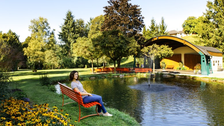 Kurpark Bad Schönau, © Niederösterreich Werbung/Doris Schwarz-König Frau sitzt auf einer roten Bank am Teich im Kurpark Bad Schönau mit Bäumen und Pavillon im Hintergrund.