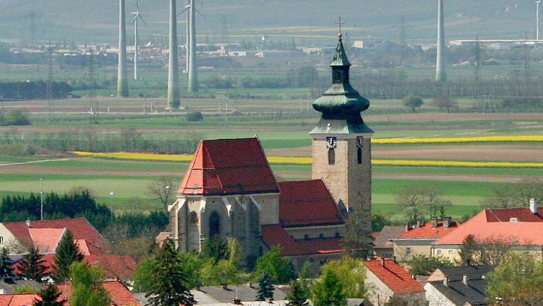 Kirche Pillichsdorf, © Thomas Falch Kirche in Pillichsdorf mit umliegenden Häusern und Windrädern im Hintergrund.