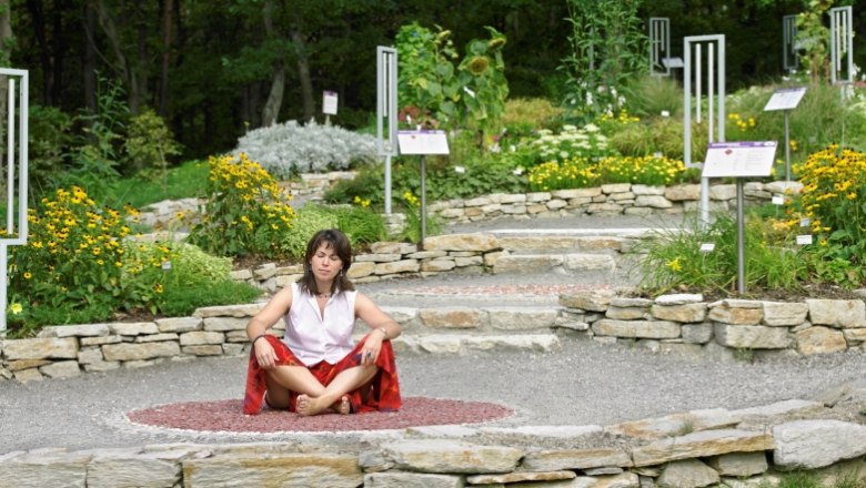 amethyst_welt_maissau_chakrenweg_ausschn_c_natur-im-garten-alexander-haiden, © Natur im Garten/Alexander Haiden Frau meditiert im Garten auf einem Steinkreis, umgeben von Blumen und Schildern.