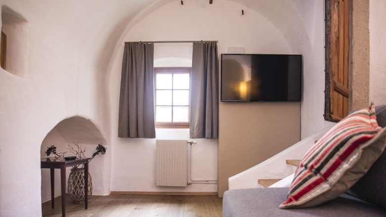 Residenz Ap. 3, © Daniela Matejschek Innenansicht eines gemütlichen Zimmers mit gewölbter Decke, Fenster mit Vorhängen, Flachbildfernseher und Sofa mit gestreiften Kissen.