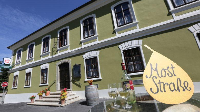 Berndl's Landgasthaus, © schwarz-koenig.at Grünes Gebäude mit weißen Fenstern, davor ein Tisch mit Weinflasche und Gläsern, Schild mit Aufschrift 'Most Straße'.