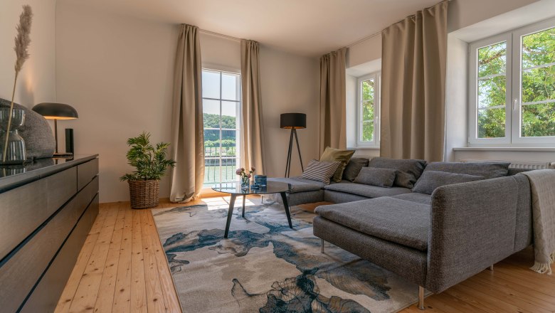 Altes Postamt Wohnzimmer-Beispiel, © Aumüller Caroline Modernes Wohnzimmer mit grauem Sofa, Holzboden und großem Fenster mit Blick ins Grüne.