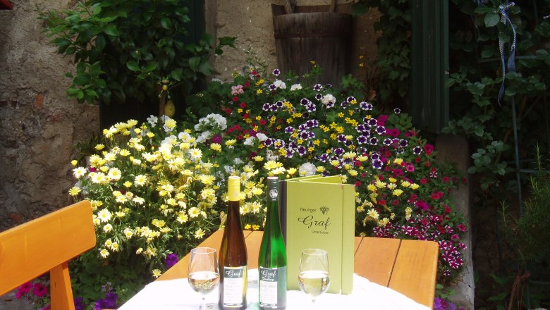 Gedeckter Tisch im Gastgarten, © Familie Graf Ein Tisch im Freien mit Weinflaschen, Gläsern und einer Speisekarte, umgeben von bunten Blumen.
