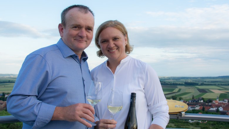 Ernst und Sonja Frischauf, © Weingut Frischauf Ein Mann und eine Frau halten Weingläser und lächeln in die Kamera, im Hintergrund eine Landschaft mit Feldern.