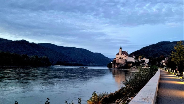 Treppelweg Richtung Schloss, © Szilágyi Ein beleuchteter Weg führt entlang eines Flusses zu einem Schloss auf einem Hügel, umgeben von bewaldeten Bergen.