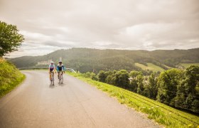 Panoramaroute bei Kirchschlag, © Wiener Alpen, Martin Fülöp Zwei Personen am Fahrrad auf einer Straße in hügeliger Landschaft mit einer Burg im Hintergrund.