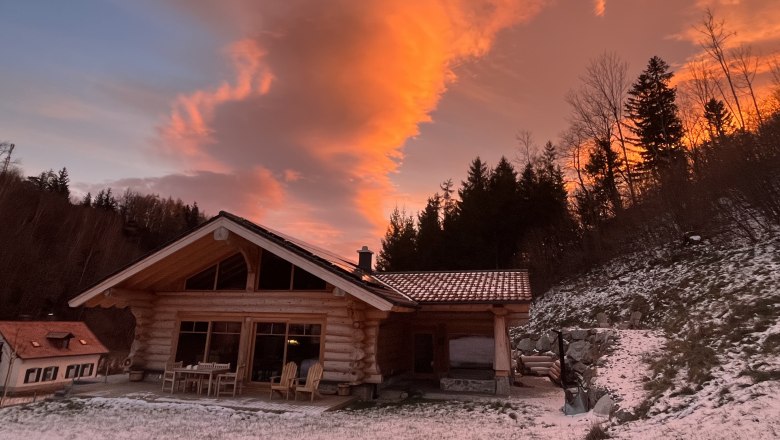 RaxChalet im Winter, © RaxChalet Ein Holzchalet im Schnee bei Sonnenuntergang mit orangefarbenem Himmel und Bäumen im Hintergrund.