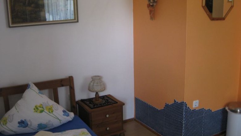 Zimmer2, © Zelenka Ein kleines Zimmer mit einem Einzelbett, Nachttisch, Lampe und orangefarbener Wand mit Mosaikfliesen.