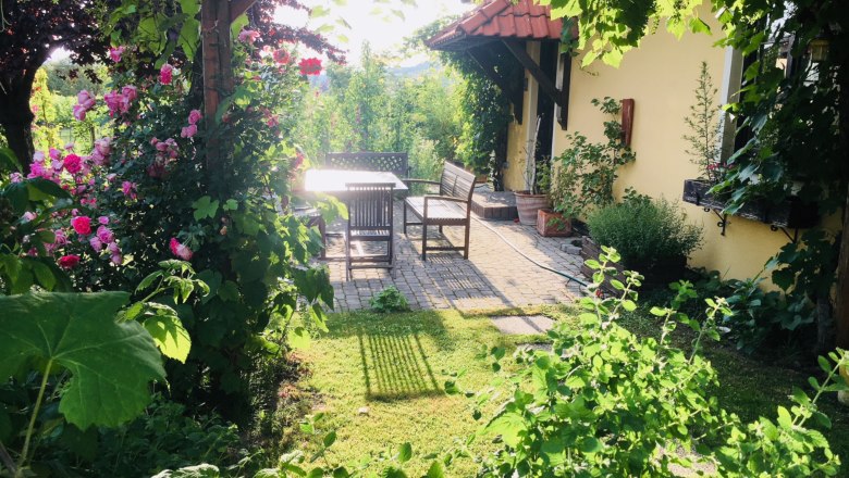 Naturgarten, © Fam. Ritz Ein idyllischer Garten mit blühenden Rosen, einem Tisch und Stühlen auf einer Terrasse neben einem gelben Haus.