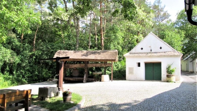 Weinviertler Kellergassenkulinarium, © D`Raschalaer Köllamauna Ein gepflasterter Hof mit einem kleinen weißen Gebäude und einem überdachten Holzgestell, umgeben von Bäumen.