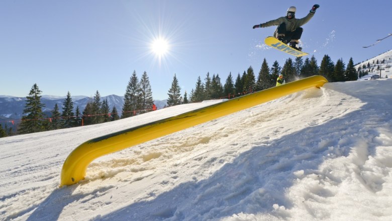 Snowpark Gemeindealpe, © Bergbahnen Mitterbach Snowboarder springt über ein gelbes Hindernis im Snowpark Gemeindealpe bei Sonnenschein.