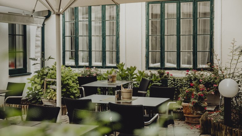 Romantische Terrasse, © Niederösterreich Werbung/Sophie Menegaldo Eine gemütliche Terrasse mit Tischen, Stühlen und Pflanzen, umgeben von großen Fenstern und einem Sonnenschirm.