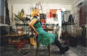 Weinviertler Fotowochen, © FLUSS / John Hilliard Eine Frau in einem grünen Kleid sitzt in einem überfüllten Raum auf einem Stuhl.