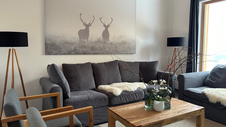 Ferienhaus Orthof - Wohnzimmer, © Ferienhäuser Gruber-List Wohnzimmer mit grauen Sofas, Holztisch und Hirschbild an der Wand.