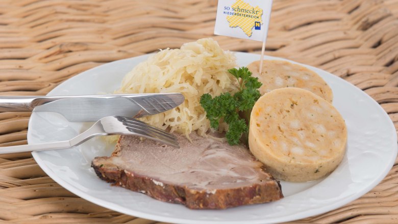 Köstliches aus der Heurigenküche, © Genussbauernhof Böhm Weißer Teller auf einem Korbtisch mit Schweinebraten, Knödeln und Sauerkraut, garniert mit Petersilie.