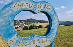 Wetterlehrpfad in Hollenthon, © Wiener Alpen Blick durch ein blaues Holzschild mit der Aufschrift 'Sonnige Grüße aus Hollenthon' auf eine ländliche Landschaft mit Hügeln und Häusern.