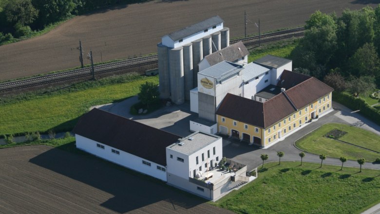 Rosenfellner Mühle, © Rosenfellner Mühle Luftaufnahme der Rosenfellner Mühle mit Gebäuden und Silos, umgeben von Feldern und Bahngleisen.
