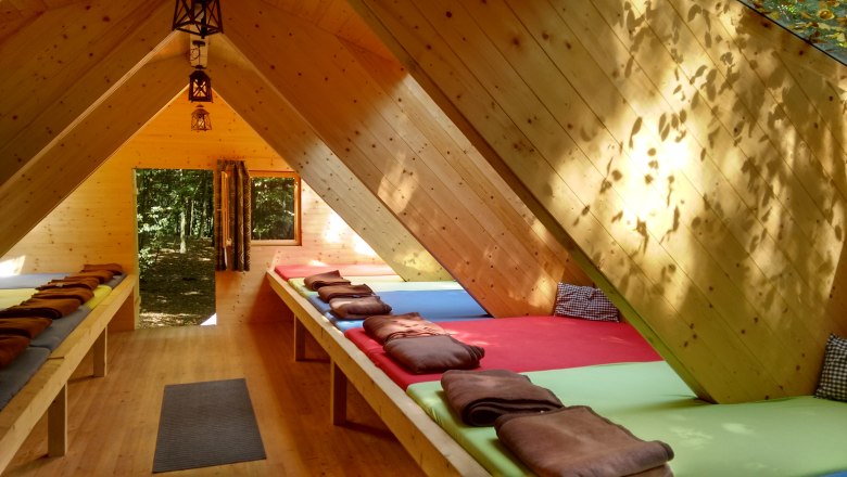 Waldcamp, © Ochys Innenansicht eines Holzhütten-Schlafsaals mit Matratzen und Decken.