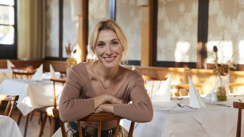 Mag. Katharina Bruckberger, © Niederösterreich Werbung/Andreas Hofer Frau sitzt lächelnd in einem Restaurant mit gedeckten Tischen.