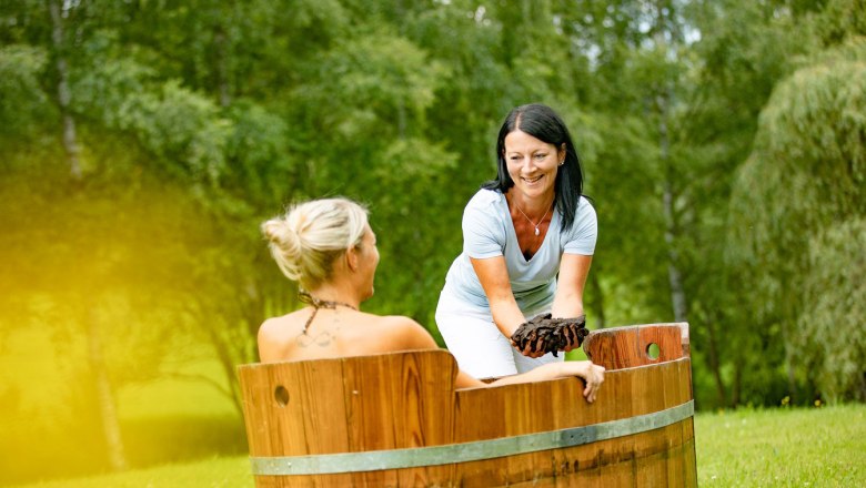 Moorwanne, © Schwarz-König/Sinzinger – www.sksfoto.at Zwei Frauen bei einer Moorbehandlung im Freien, eine sitzt in einer Holzwanne, die andere reicht ihr Moor.