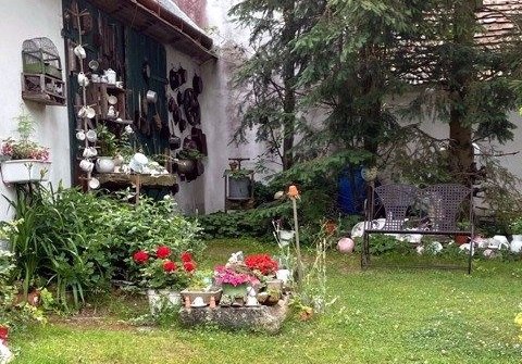 Nostalgieschaugarten, © Erika Pascher Ein Garten mit alten Töpfen und Dekorationen an einer Wand, Blumenbeet und zwei Stühlen.