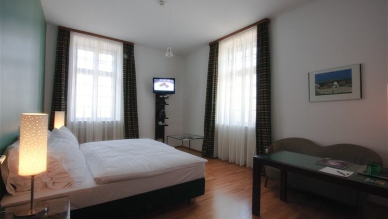 Zimmer Schloss Grafenegg, © Mörwald GmbH Ein Hotelzimmer mit Doppelbett, zwei Fenstern, einem kleinen Fernseher und einem Tisch mit Stuhl.