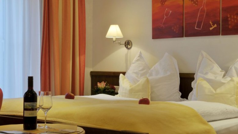 Zimmerbeispiel im Weinhof Aufreiter, © Steve Haider Gemütliches Hotelzimmer mit Doppelbett, gelber Bettdecke, Weinflasche und Gläsern auf Tisch, Wandkunst mit Weinmotiv.