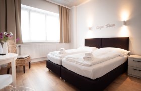 Doppelzimmer, © Doris Eminger Modernes Doppelzimmer mit zwei Betten, weißen Bettdecken und Handtüchern. Schriftzug 'Carpe Diem' an der Wand, helle Vorhänge und Holzboden.